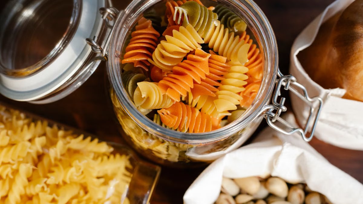 From above glass jar with colorful rotini placed on table near fusilli in container and pistachios and bread in bags